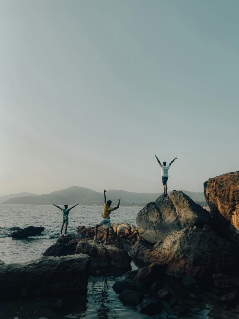 Three friends celebrating on rocky seacoast in Goa, India with arms raised, embracing adventure.