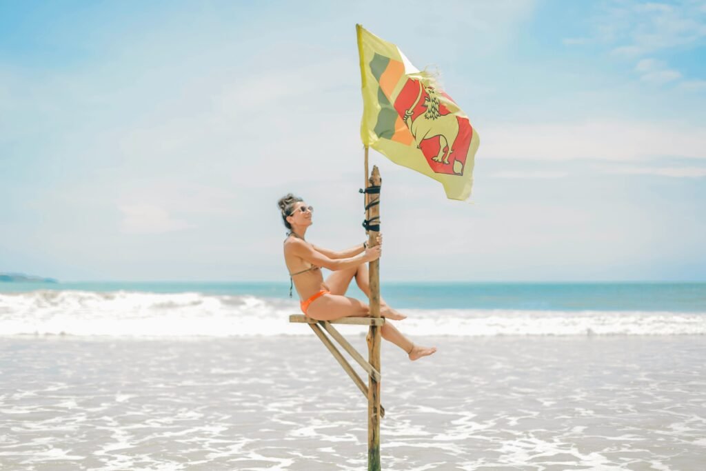 Woman in a bikini sitting on a wooden structure by the ocean with a Sri Lankan flag waving in the breeze.