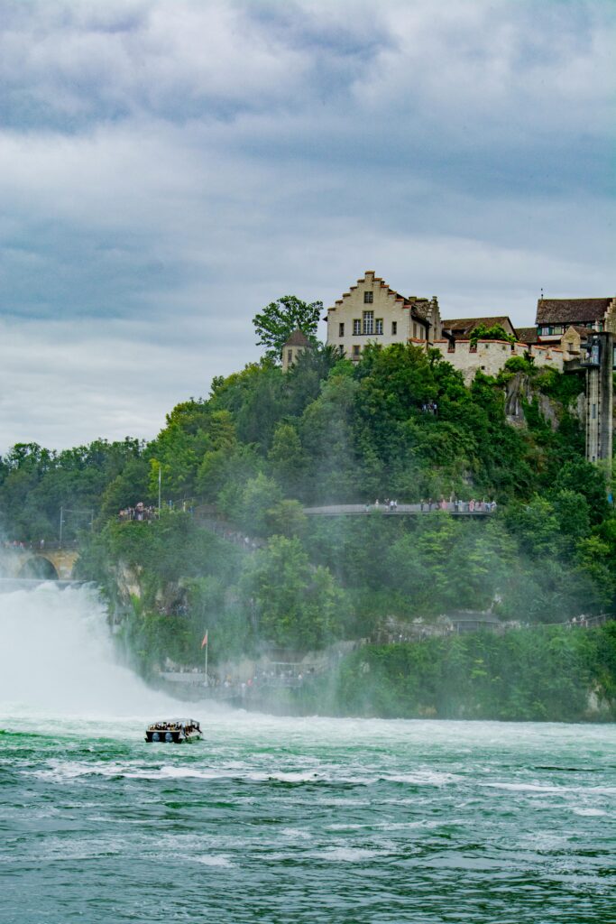 pexels-photo-27303387-27303387 Historic castle overlooking the majestic Rhine Falls in Switzerland, with lush greenery and a boat on the water.